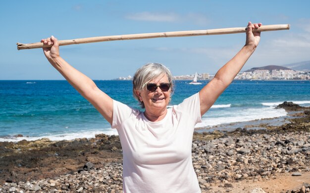 Vista frontale di un popolo, donna anziana che esegue alcuni esercizi per le spalle sulla spiaggia. Orizzonte sull'acqua. Cielo azzurro e mare sullo sfondo. Vacanze o pensione. Serenità e relax