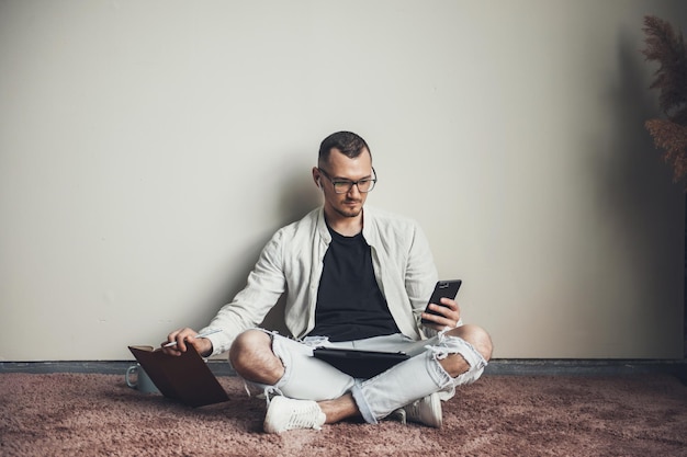 Vista frontale di un uomo caucasico seduto per terra che guarda lo smartphone mentre tiene un libro mo...