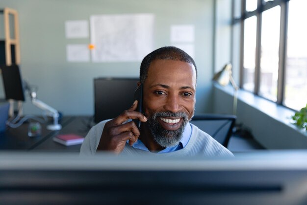 Vista frontale di un uomo d'affari afroamericano che lavora nell'ufficio moderno, seduto accanto a una scrivania, sorridente, parlando sul suo smartphone e utilizzando un computer desktop. Distanziamento sociale e autoisolamento in