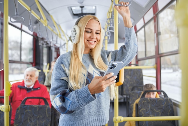 Vista frontale di una bella ragazza bionda che usa il telefono sorridente in piedi all'interno di un autobus in movimento raccolto di donna