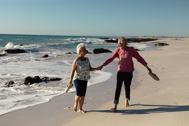 Vista frontale di una coppia caucasica anziana in spiaggia al sole, tenendo le scarpe e camminando a piedi nudi tenendosi per mano, sorridendo e parlando, con mare e cielo blu sullo sfondo