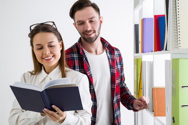 Vista frontale di una coppia di studenti in una biblioteca. La ragazza tiene in mano un libro, il ragazzo sta leggendo da sopra la sua spalla