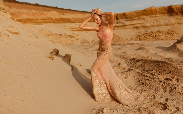 Vista frontale di una donna bionda in splendido abito d'oro in posa nel deserto, su sfondo di carriera di sabbia.