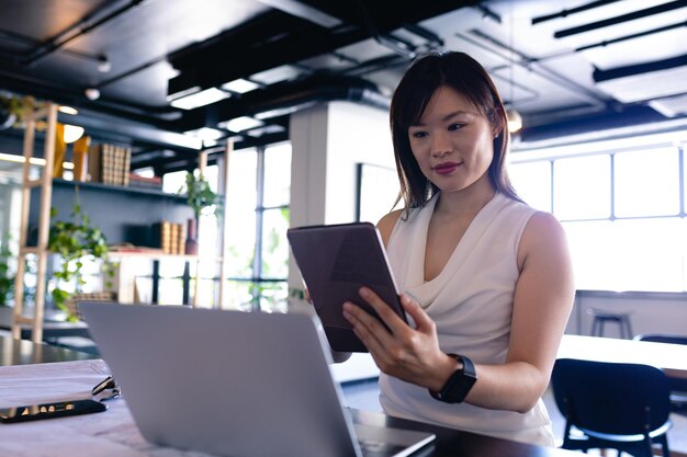 Vista frontale di una donna d'affari asiatica che indossa abiti eleganti, lavora in un ufficio moderno, sta accanto a una scrivania e usa il suo computer portatile e tablet.