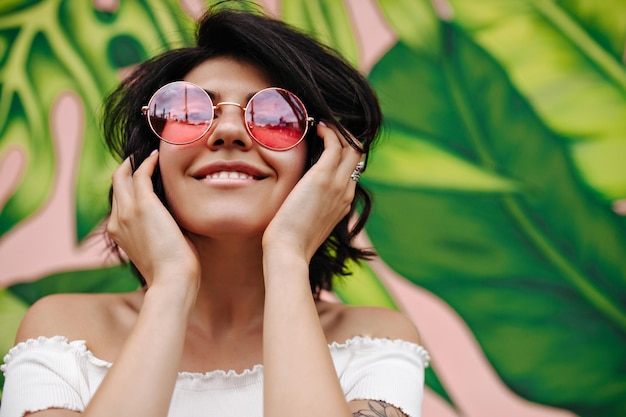 Vista frontale di una ragazza straordinaria con occhiali rotondi rosa Modello femminile ridente in posa vicino a graffiti verdi