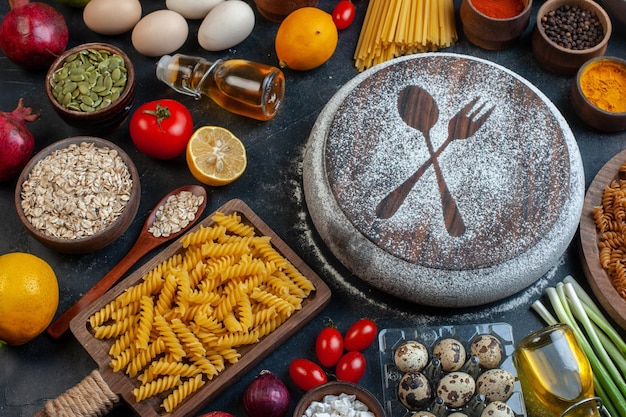 Vista frontale farina bianca a forma di cucchiaio e forchetta intorno alle uova pomodori condimenti e pasta su sfondo scuro colore pasta verdure pasto cibo