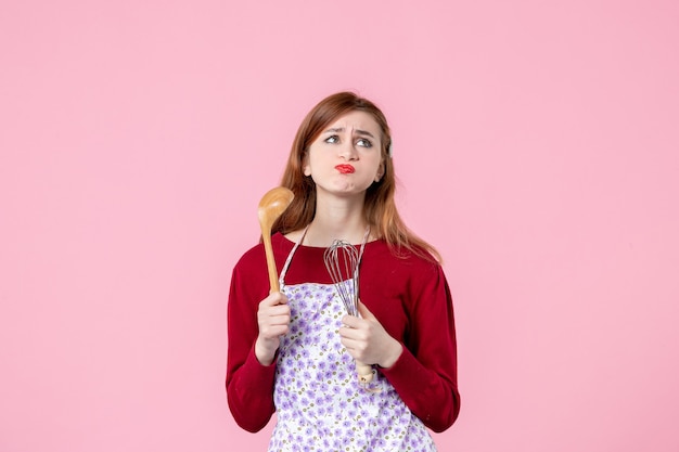 vista frontale giovane casalinga che tiene frusta e cucchiaio di legno su sfondo rosa cucina cucina donna torta torta uniforme professione pasta