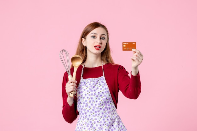 Vista frontale giovane casalinga che tiene la frusta e la carta bancaria su sfondo rosa cucina torta torta donna colore shopping cucina denaro cibo