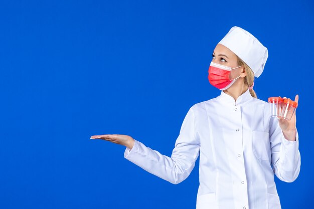 vista frontale giovane infermiera in tuta medica con maschera rossa che tiene flaconi su sfondo blu medico sanitario covid- pandemia medicina ospedaliera