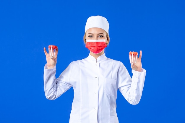 vista frontale giovane infermiera in tuta medica con maschera rossa che tiene flaconi su sfondo blu virus sanitario pandemia covid- medicina della droga ospedaliera