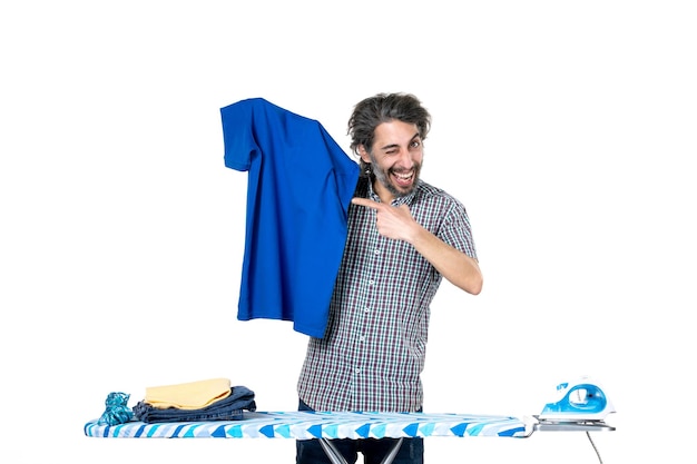 vista frontale giovane maschio dietro asse da stiro pieghevole camicia blu su sfondo bianco lavori domestici ferro da stiro lavanderia uomo vestiti a casa machine