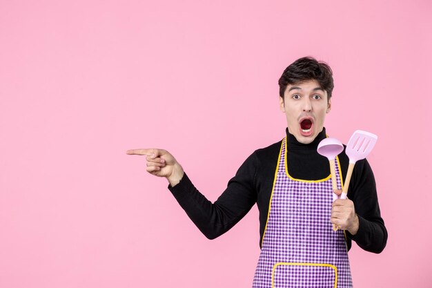 Vista frontale giovane maschio in mantello che tiene cucchiai rosa su sfondo rosa pasta professione cucina uniforme da lavoro colori cucina orizzontale