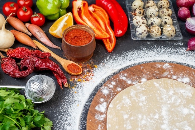 Vista frontale ravvicinata dell'impasto circolare su una tavola di legno con farina intorno e spezie di verdure fresche su superficie nera