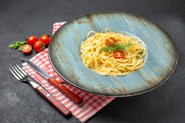 Vista frontale ravvicinata di deliziosi spaghetti serviti con pomodori verdi e posate su un asciugamano spogliato rosso su sfondo nero