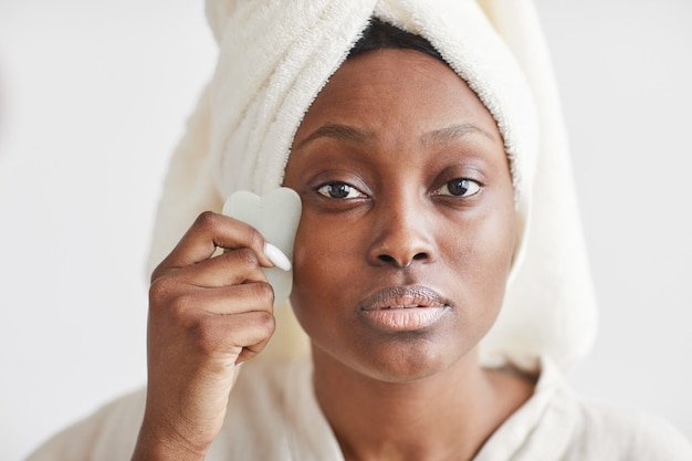Vista frontale ritratto di bella donna afro-americana che fa un massaggio al viso mentre si gode la routine di cura della pelle a casa e guarda la macchina fotografica, copia spazio