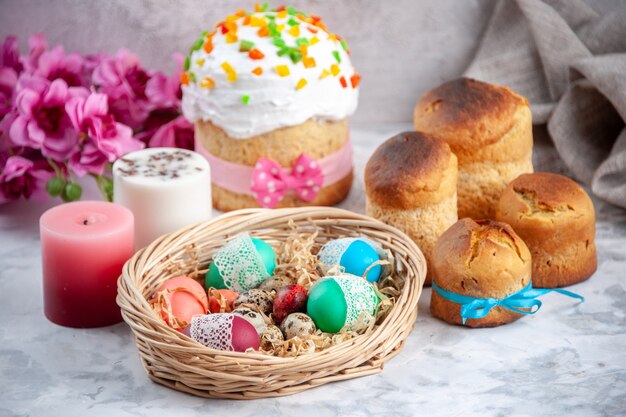 vista frontale uova di pasqua colorate all'interno del cesto con candele e torta su superficie bianca