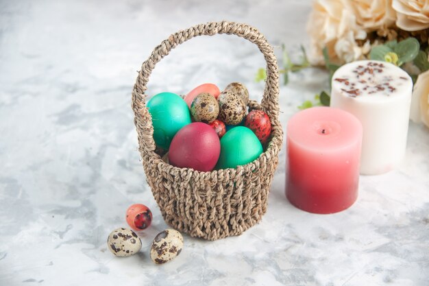 vista frontale uova di pasqua colorate all'interno del cesto progettato con fiori e candele su superficie bianca