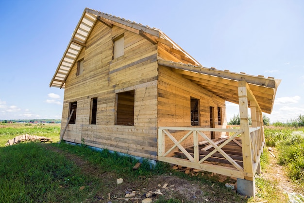 Vista grandangolare del cottage tradizionale ecologico di legno non finito dei materiali naturali del legname con il tetto ripido e il portico in costruzione in vicinanza verde sul fondo dello spazio della copia del cielo blu.