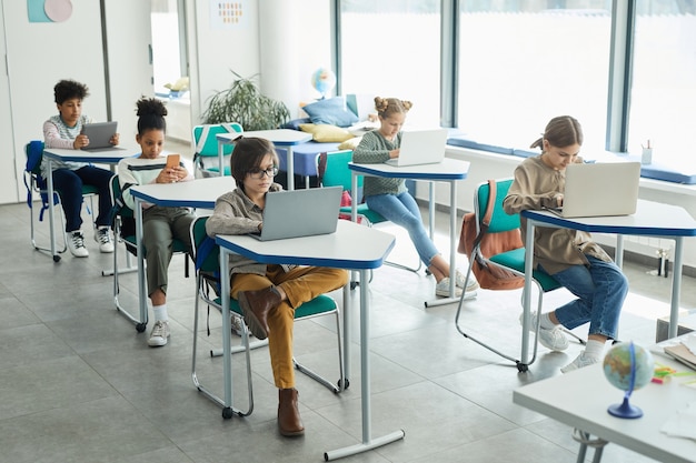 Vista grandangolare su un gruppo eterogeneo di bambini piccoli che usano gadget mentre sono seduti ai banchi in aula scolastica, copia spazio