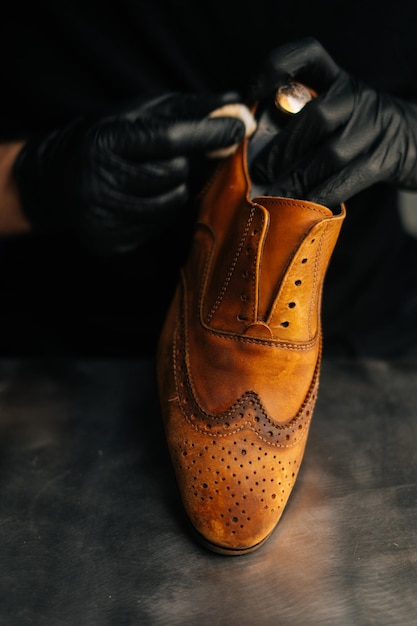 Vista ingrandita del calzolaio che indossa guanti di lattice neri che puliscono le vecchie scarpe in pelle marrone chiaro con uno straccio per un successivo restauro