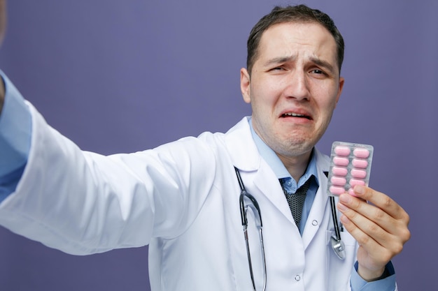 Vista ingrandita del giovane medico maschio spaventato che indossa accappatoio medico e stetoscopio intorno al collo guardando la fotocamera piangendo allungando la mano verso la fotocamera che mostra la confezione di capsule prendendo selfie