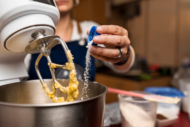 Vista ingrandita della mano di un cuoco che mette una polvere bianca in una ciotola del mixer per la preparazione della ricetta fatta in casa di alfajores argentini