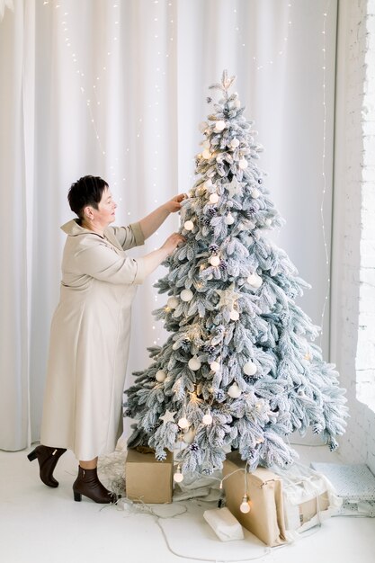 Vista laterale a tutta lunghezza di gioiosa donna matura che indossa un elegante abito lungo che decora il bellissimo albero di Natale