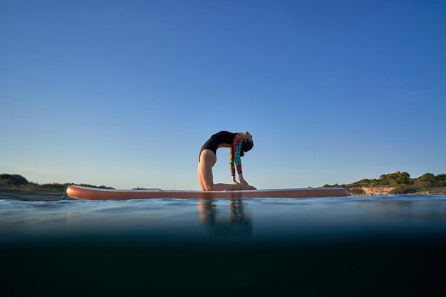 Vista laterale a tutta lunghezza di una donna in forma irriconoscibile in costume da bagno in piedi sulla tavola SUP e facendo posa yoga Ushtrasana in serata in mare