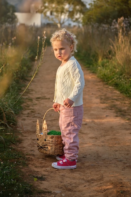 Vista laterale adorabile bambina in abiti eleganti che trasportano cesto di vimini e in piedi sulla stretta strada rurale nella natura verdeggiante