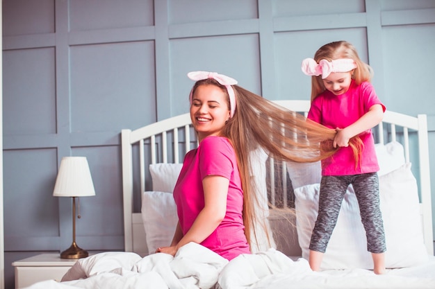 Vista laterale bambina in pinktshirt in piedi sul letto e pettinare sua madre Giovane bella donna seduta a letto mentre sua figlia gioca con i suoi capelli lunghi Salone di bellezza a casa