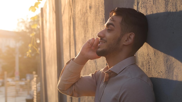 Vista laterale bello barbuto arabo ispanico uomo d'affari cliente ragazzo in piedi vicino al muro di pietra che parla al cellulare rispondendo ai colloqui di chiamata con la conversazione dello smartphone utilizzando il cellulare wireless in città