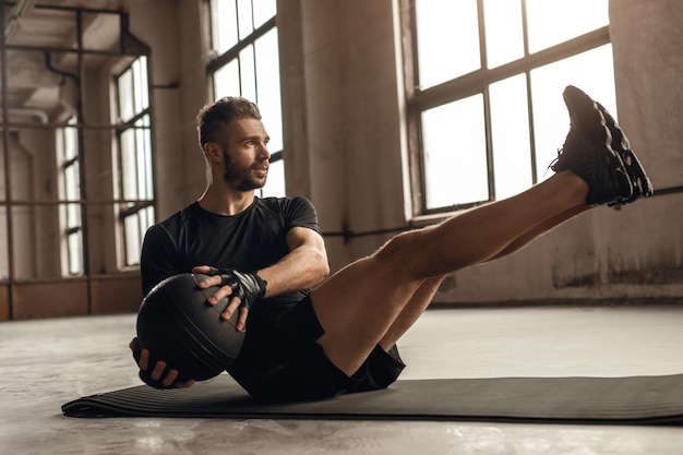 Vista laterale completa del corpo di un forte maschio adulto barbuto in abbigliamento sportivo che fa esercizio di torsione laterale seduto con palla medica durante l'allenamento degli addominali in palestra