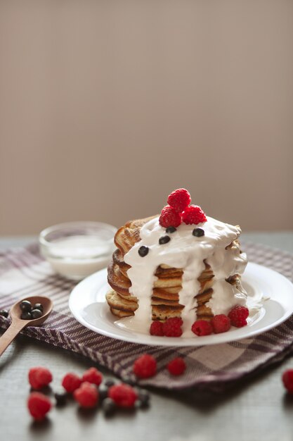 Vista laterale dei pancake saporiti nello stack con panna acida e le bacche sul piatto bianco