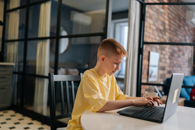 Vista laterale del bel ragazzo di 10 anni in maglietta gialla che digita sulla tastiera del computer portatile durante la lezione online che studia a distanza Scolaro bambino focalizzato che utilizza il computer