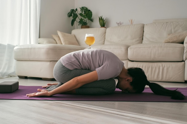 Vista laterale del corpo completo di una donna pacifica che pratica la postura di Balasana con un bicchiere di birra sulla schiena durante una sessione di yoga in camera