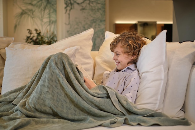 Vista laterale del corpo pieno di un bambino felice che legge un libro interessante mentre giace su un comodo divano sotto una coperta in un luminoso soggiorno a casa