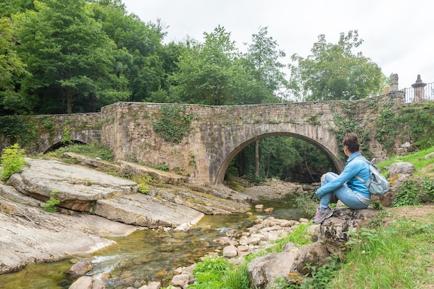Vista laterale del corpo pieno di una donna irriconoscibile seduta su pietre e ammirando la vista del vecchio ponte ad arco in pietra sul torrente stretto a Barcena Mayor in Cantabria in Spagna