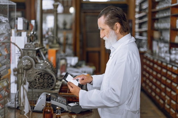 Vista laterale del farmacista uomo anziano concentrato professionale utilizzando tablet digitale che lavora in antica farmacia con interni d'epoca