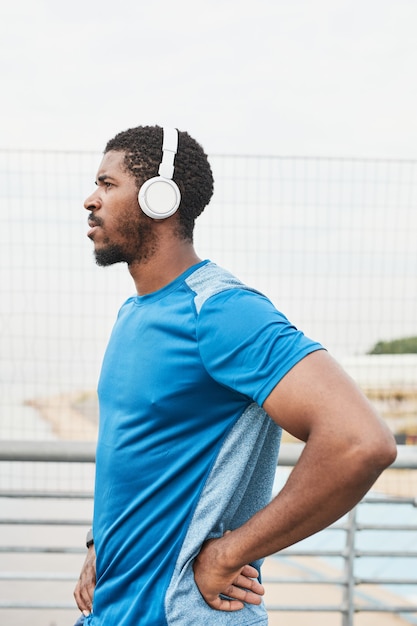 Vista laterale del giovane atleta africano in cuffie wireless che ascolta la musica durante l'allenamento all'aperto