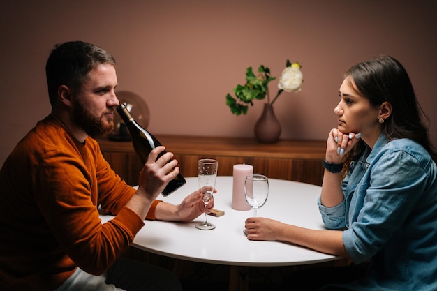 Vista laterale del giovane barbuto che annusa una bottiglia di vino durante la conversazione con una bella ragazza seduta insieme al tavolo. Coppia d'amore che celebra l'anniversario o il giorno di San Valentino con una cena romantica.