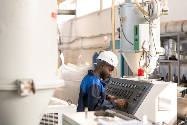 Vista laterale del giovane lavoratore africano in tuta e elmetto protettivo in piedi davanti al pannello di controllo di un'enorme macchina industriale e meccanismo di regolazione