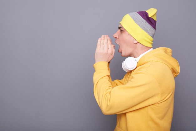 Vista laterale del giovane uomo adulto che indossa una felpa con cappuccio gialla e un cappello a cuffia in posa isolato su sfondo grigio urlando con le mani vicino alla bocca facendo annunciare urlando forte