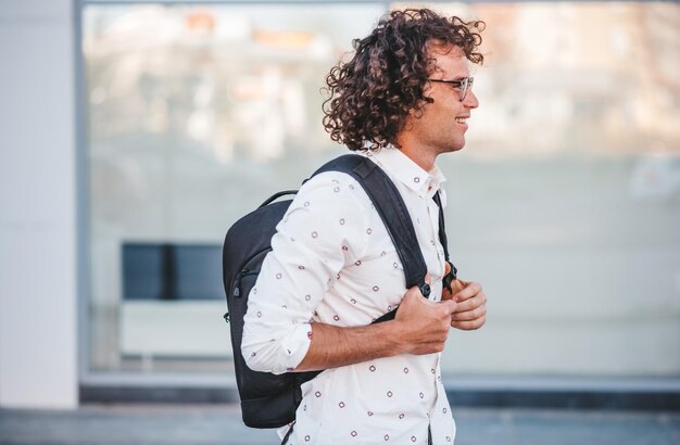 Vista laterale del giovane uomo d'affari con lo zaino sulla strada della città Il maschio caucasico con i capelli ricci che sorride e che guarda un lato in camicia casual sta andando all'università Concetto di persone e stile di vita