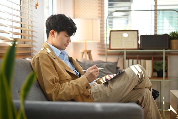 Vista laterale del libero professionista maschio asiatico che lavora online controllando la posta elettronica sul tablet mentre si è seduti in soggiorno
