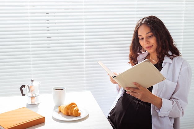 Vista laterale del libro di lettura felice della donna incinta mentre si consuma la colazione del mattino con caffè e