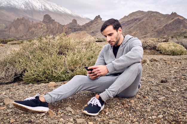 Vista laterale del maschio concentrato barbuto in messaggio di testo activewear sul telefono cellulare mentre si riposa su un terreno pietroso vicino al vulcano Teide a Tenerife nelle Isole Canarie in Spagna