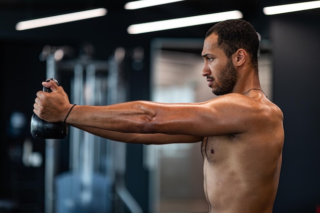 Vista laterale del muscoloso uomo nero senza camicia che si allena con kettlebell in palestra