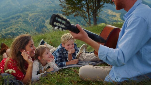 Vista laterale del padre rosso che suona la chitarra per una donna sorridente con bambini sdraiati sull'erba da vicino La famiglia allegra si diverte ad ascoltare musica sulla collina verde Genitori felici che trascorrono le vacanze estive con i bambini