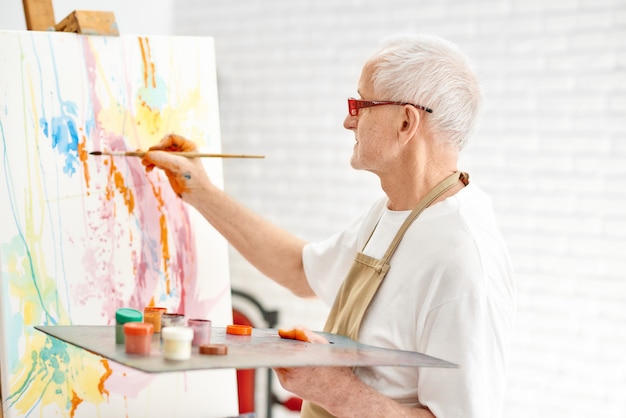 Vista laterale del pittore di talento senior mentre dipingeva il suo capolavoro in studio d&#39;arte luminosa.