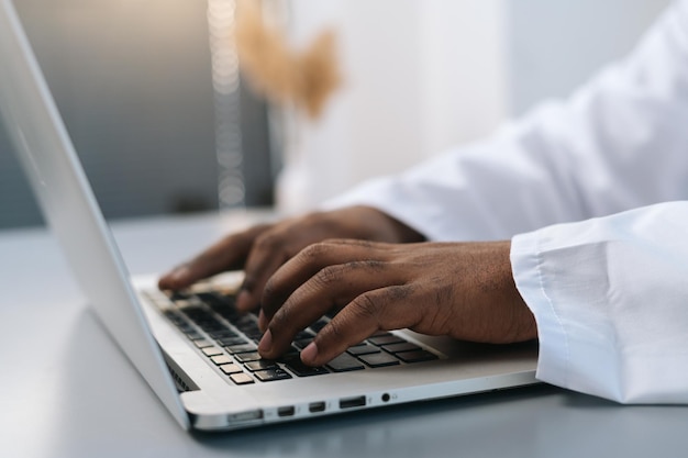 Vista laterale del primo piano le mani del medico maschio afroamericano irriconoscibile che indossa camice bianco che lavora digitando sul computer portatile